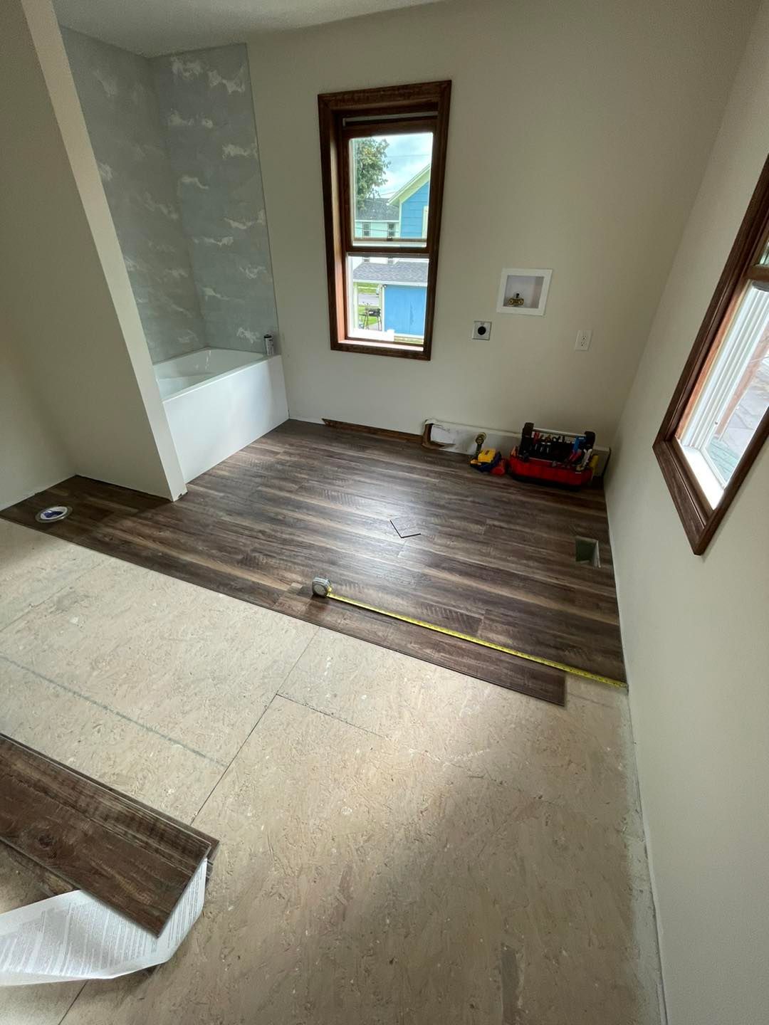 Bathroom under renovation with new wood-look flooring being installed. Light walls, tub, window, and tools.