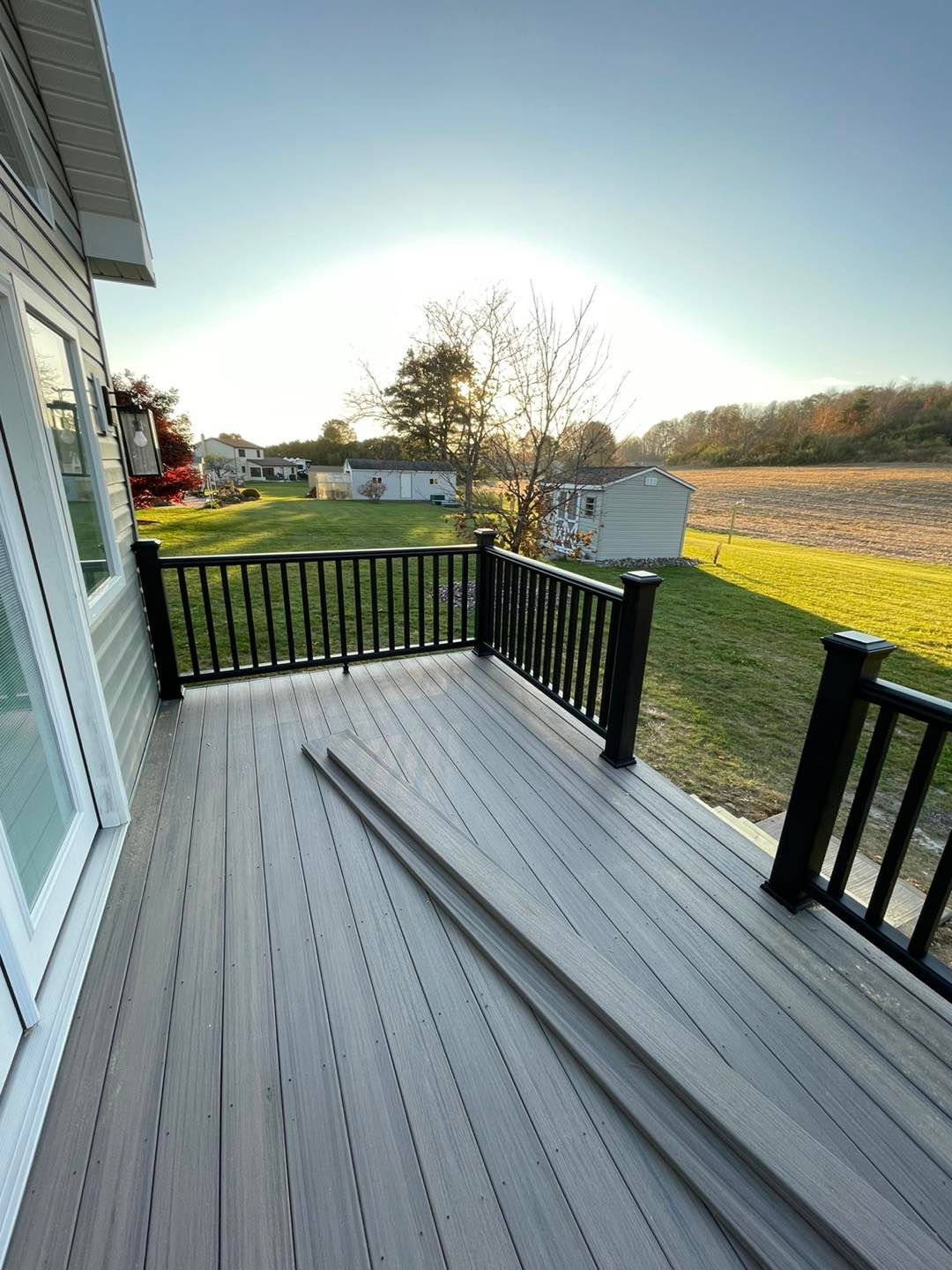 Deck with black railing, gray composite decking, overlooking a grassy field and houses under a sunny sky.