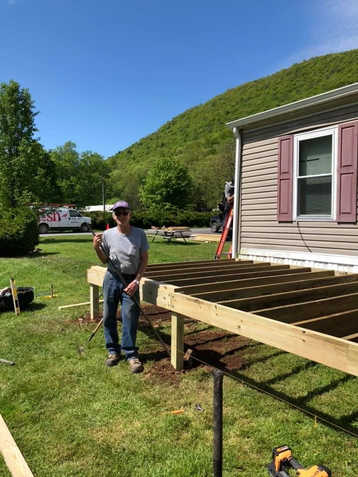 Man giving thumbs up stands on partially built wooden deck beside a house with mountain backdrop.