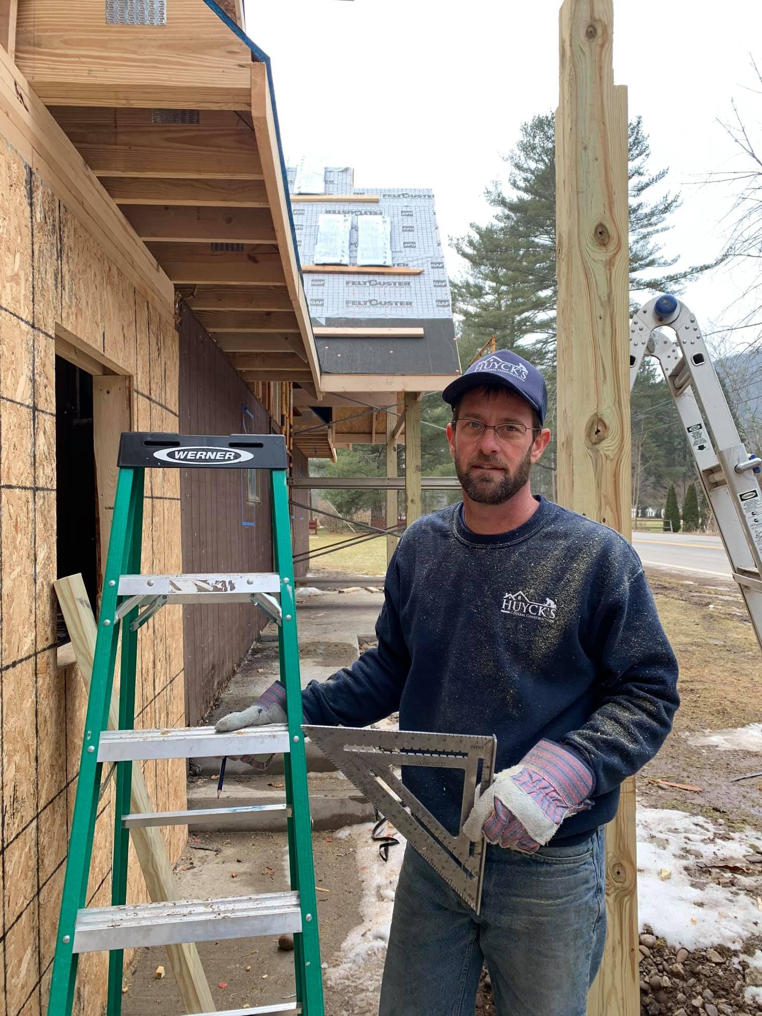 A carpenter with glasses, square tool, and ladder builds outside.