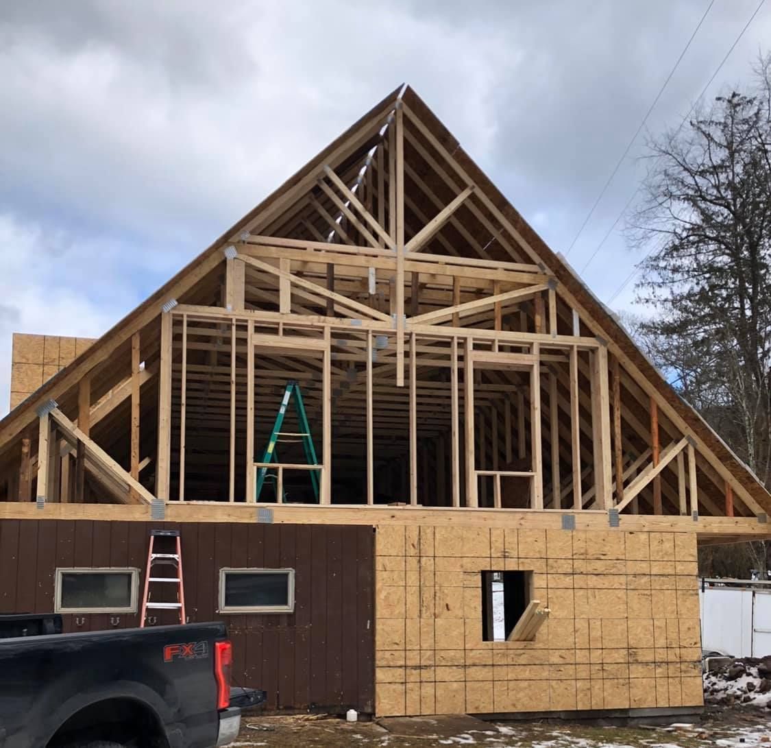 Framed wooden structure of a house under construction with a high-peaked roof, against a cloudy sky.