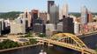 Pittsburgh skyline with yellow bridge over a river, tall buildings against a blue sky.