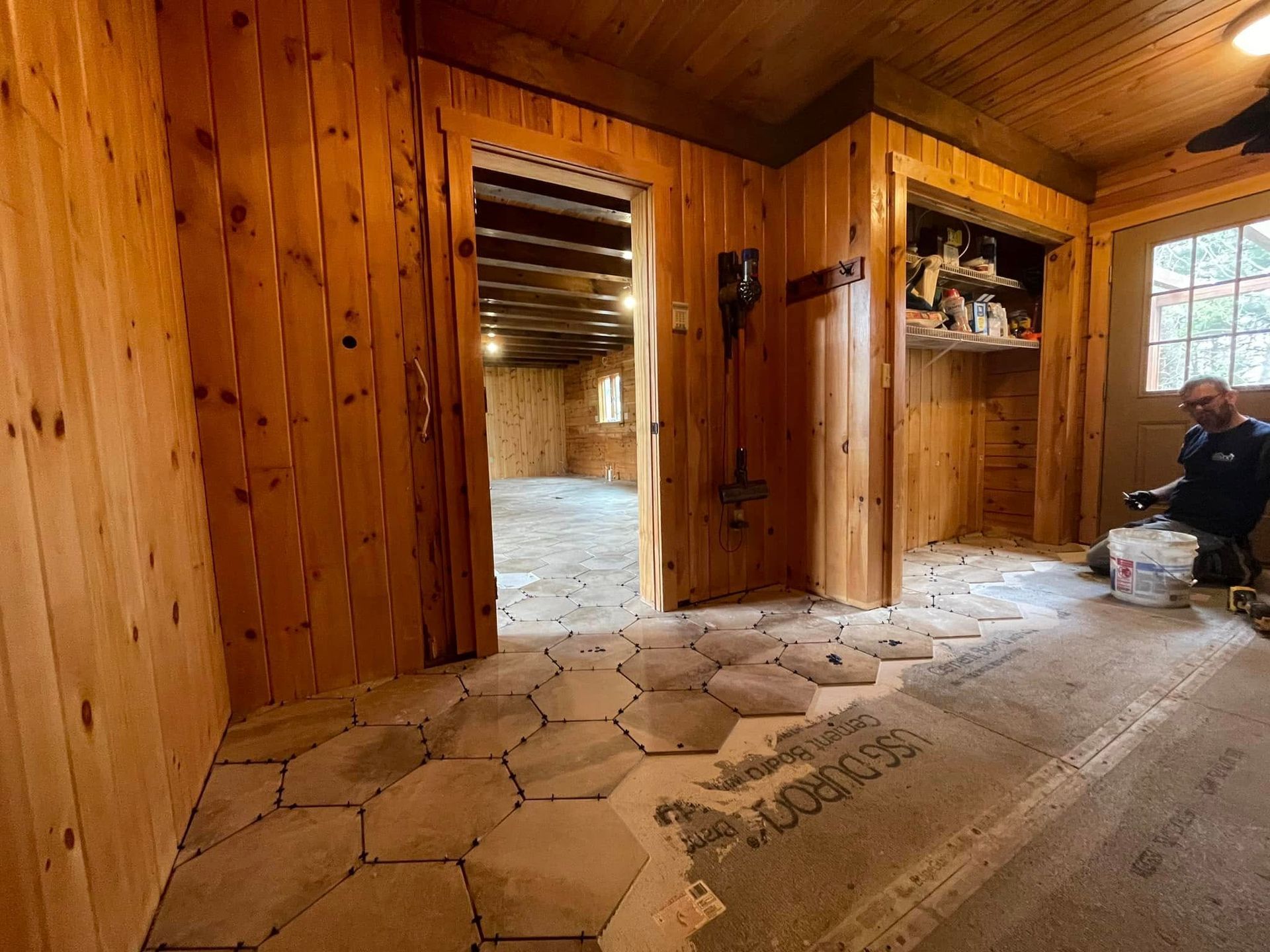 Man tiling a room with wood paneling and hexagonal stone tiles.