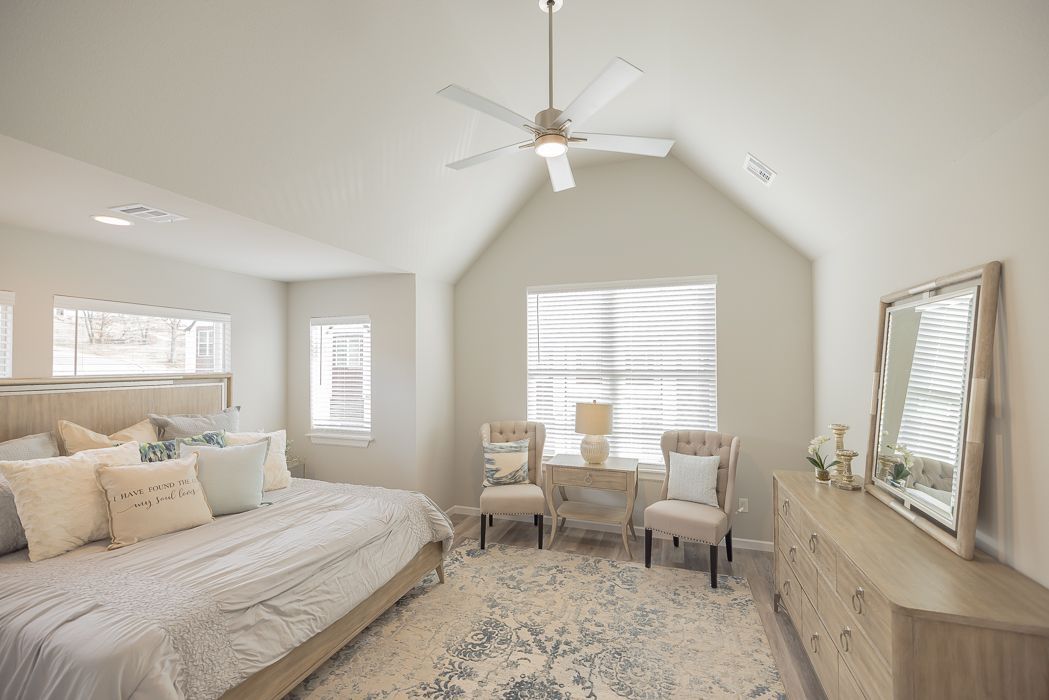 Bright bedroom with king bed, vaulted ceiling, windows, dresser, rug, and sitting area.