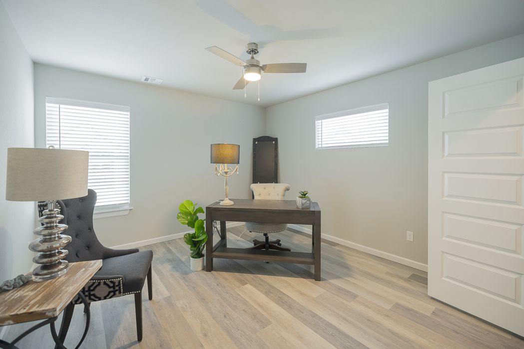 Home office with desk, chair, lamp, and window blinds; light gray walls and wood flooring.