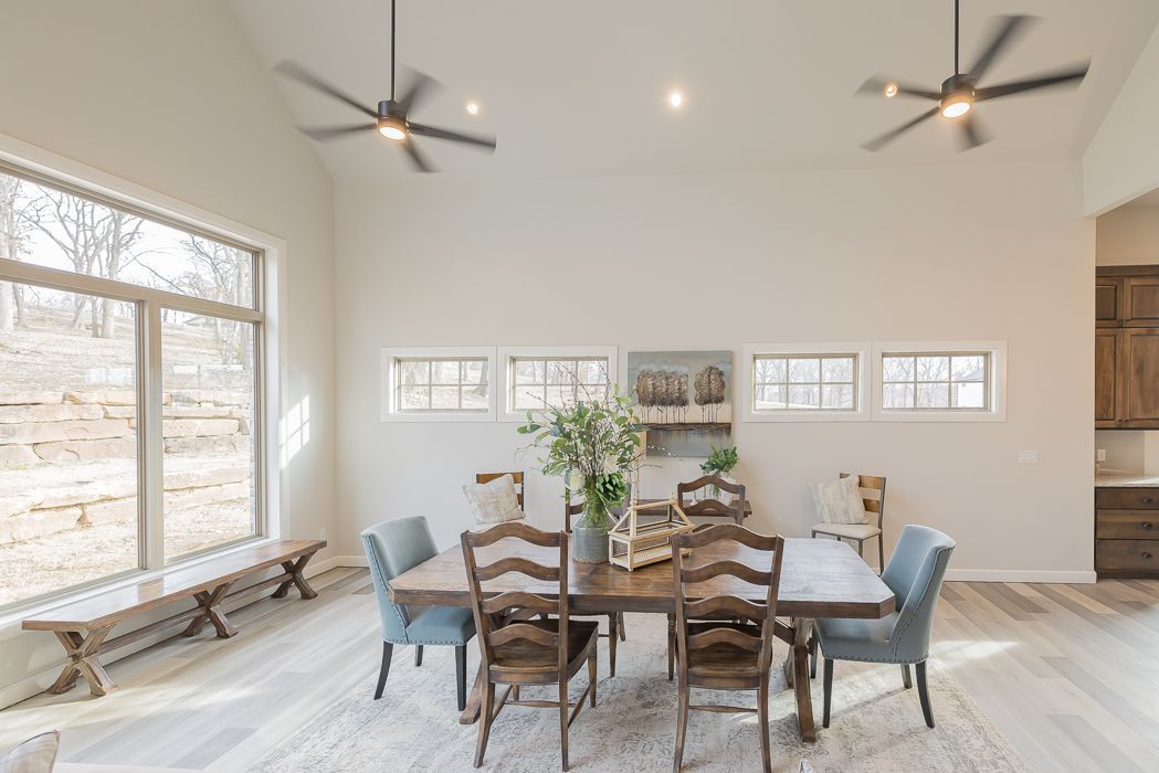 Dining room with table and chairs; large windows, high ceiling, and neutral color palette.