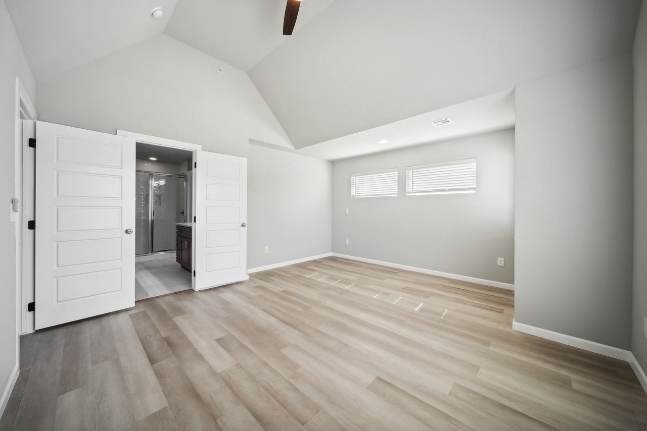Empty bedroom with gray walls, vaulted ceiling, light wood floors, and open double doors to a bathroom.