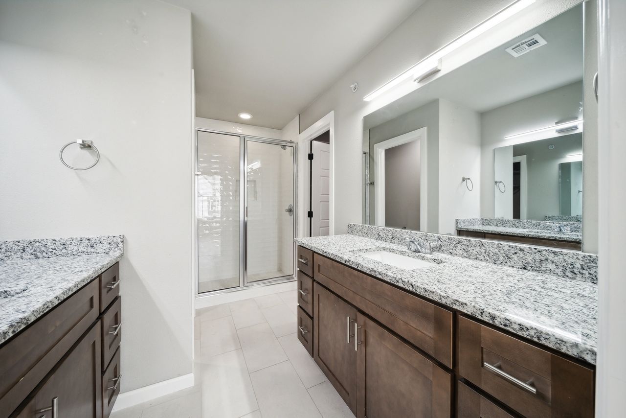 A bright, modern bathroom with a double vanity, shower, and large mirror. Gray walls and granite countertops.