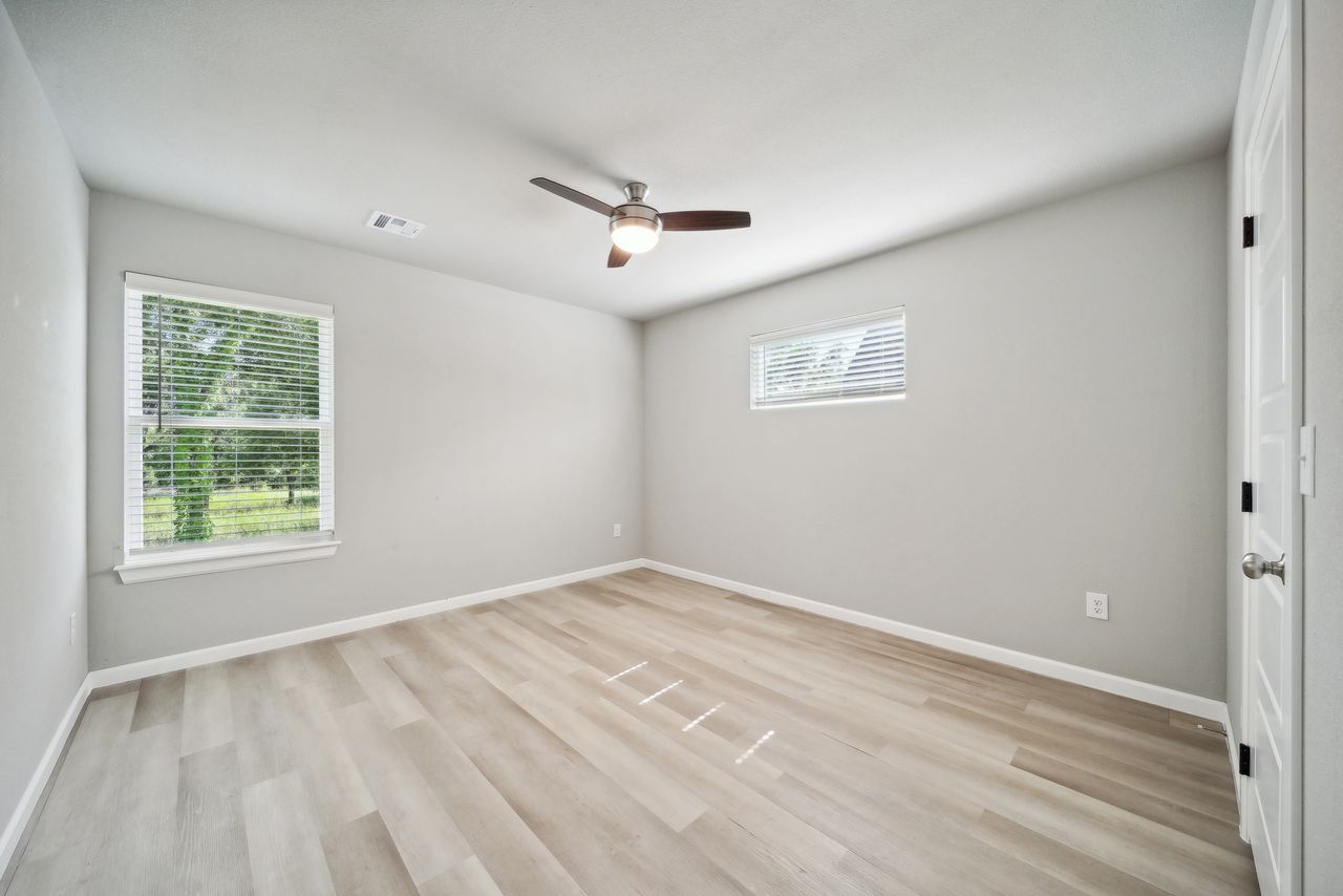 Empty room with light gray walls, wood-look floors, two windows, ceiling fan, and white door.