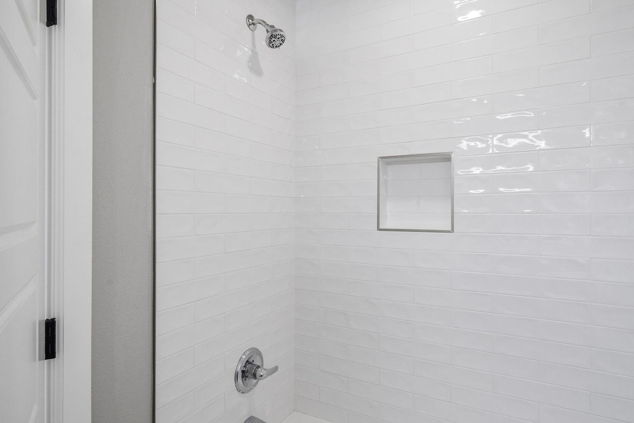 White-tiled shower with recessed shelf, chrome fixtures, and showerhead.