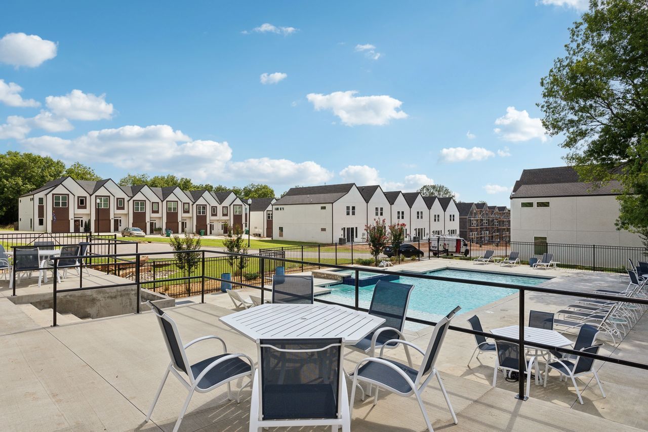 Outdoor patio with pool and modern townhouses under a blue sky. Tables, chairs, and landscaping.