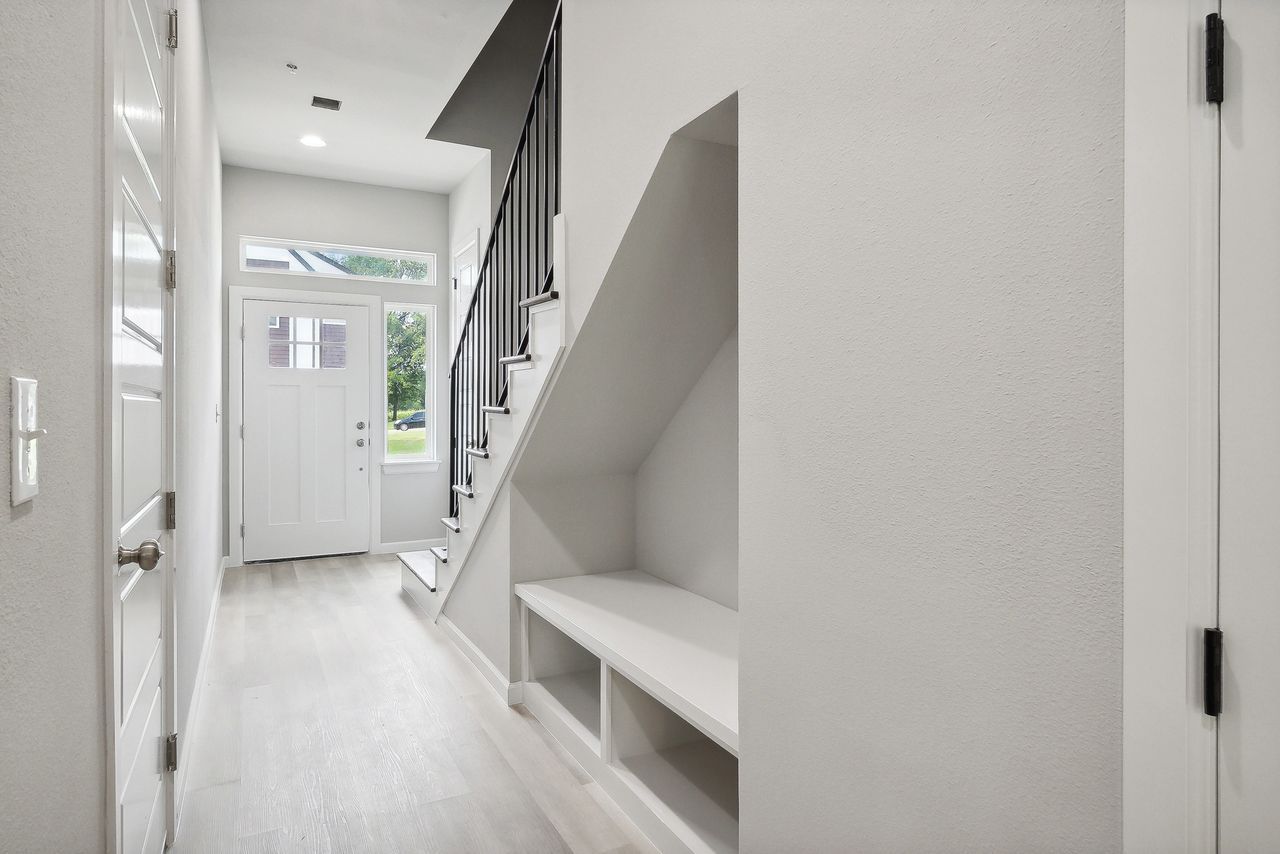 Hallway with stairs, built-in bench. White walls, light wood-look flooring, and front door visible.