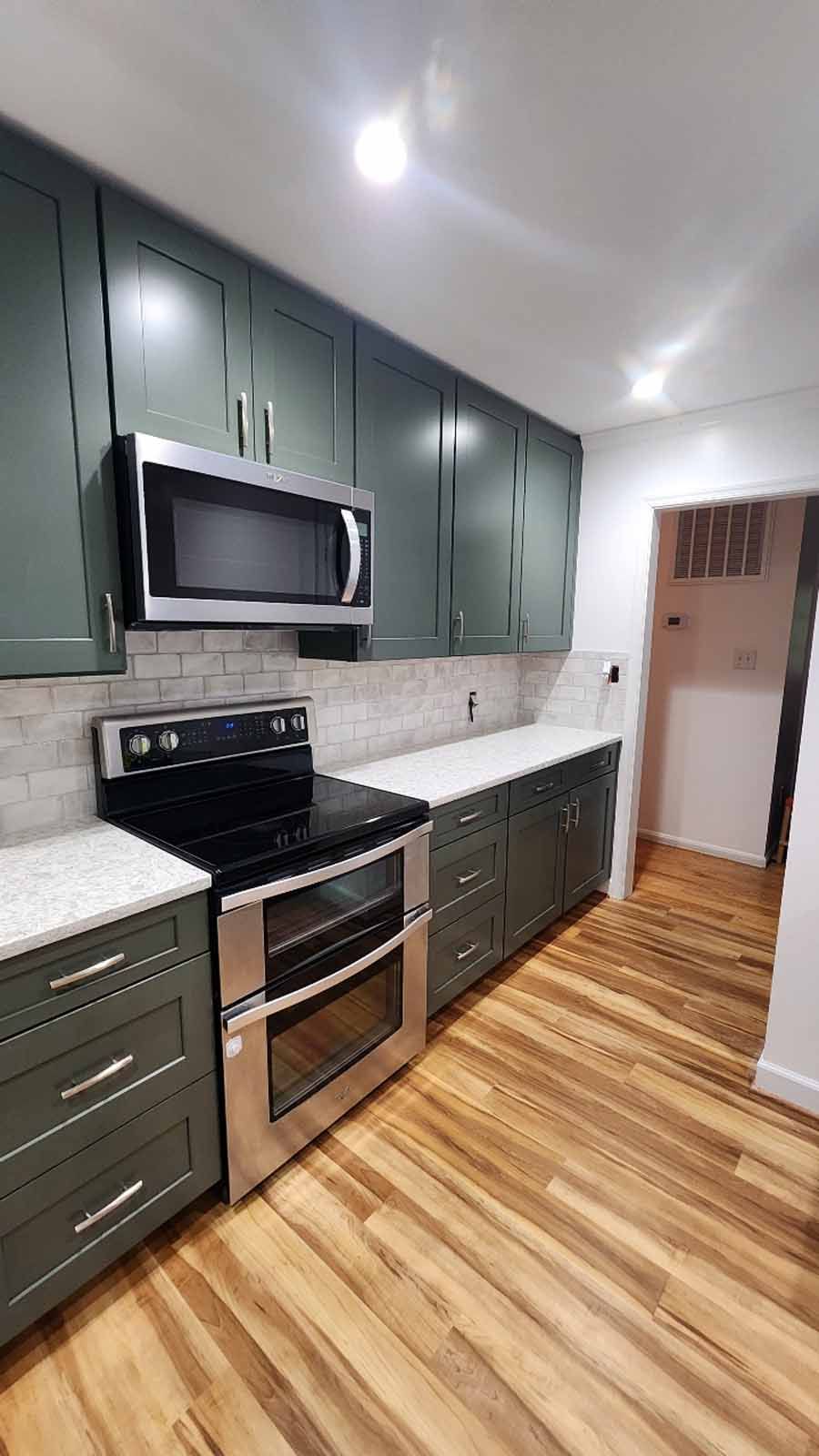 Green kitchen cabinets and stainless steel appliances. White backsplash, wooden floor.