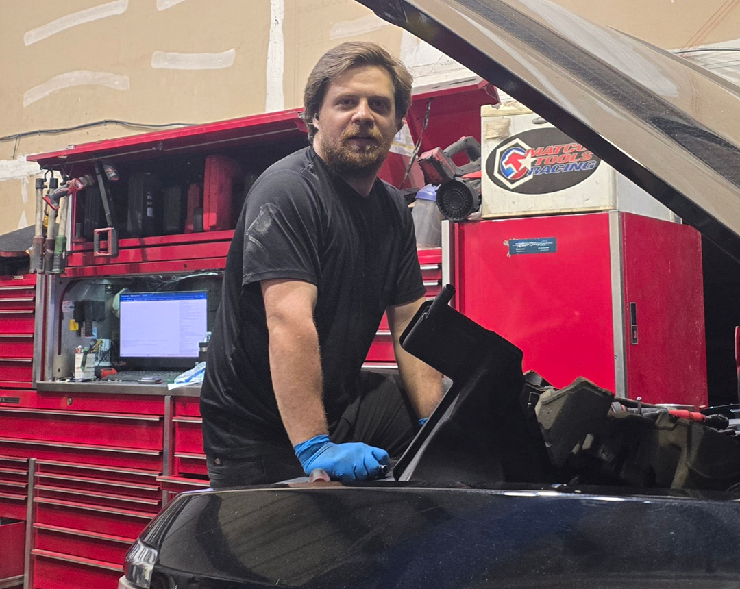 Mechanic working on a car engine, wearing gloves.  Garage setting, red tool cabinets, man smiling at camera.  | Integrity Auto Repair