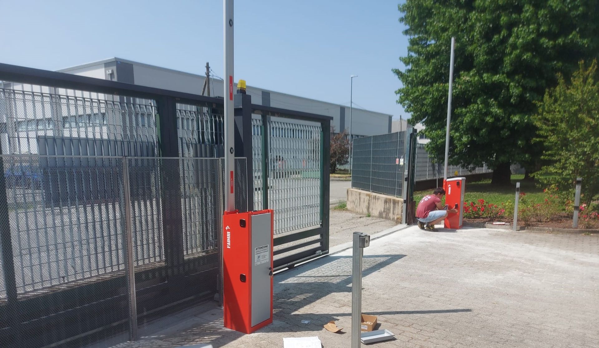 Due bracci automatici rossi e grigi all'ingresso, persona che lavora ai comandi del cancello all'esterno.