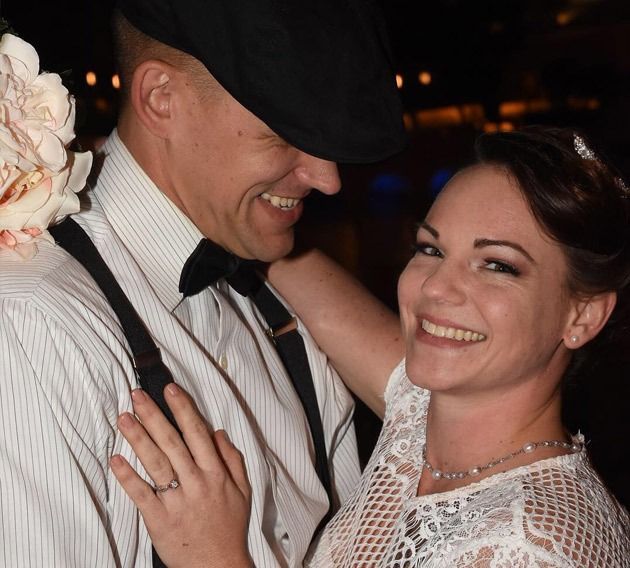 Bride and groom smiling and embracing. The groom wears a cap and suspenders. The bride wears a white lace dress.