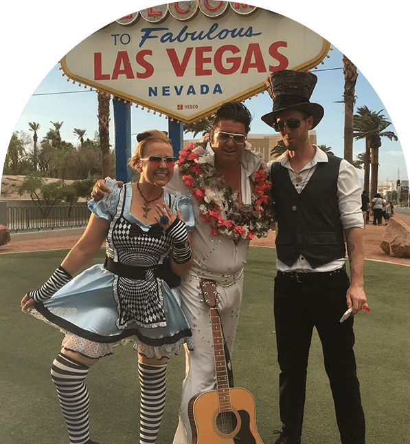 Three people pose in front of the Las Vegas sign: Alice in Wonderland, Elvis, and a man in steampunk attire.
