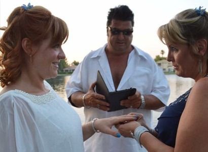Two women exchanging rings during an outdoor wedding ceremony, officiated by a man in sunglasses.
