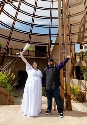 Couple celebrating: woman in white dress raises bouquet, man in black vest raises fist, outside near dome.