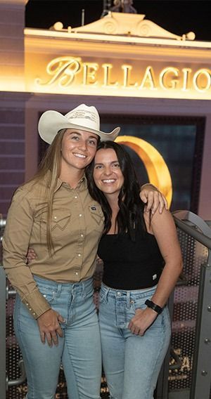 Two women smiling, posing in front of the Bellagio sign in Las Vegas. One wears a cowboy hat, the other a black tank top.