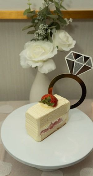 Cake slice topped with strawberry and diamond ring cake topper on white plate. Flowers in background.