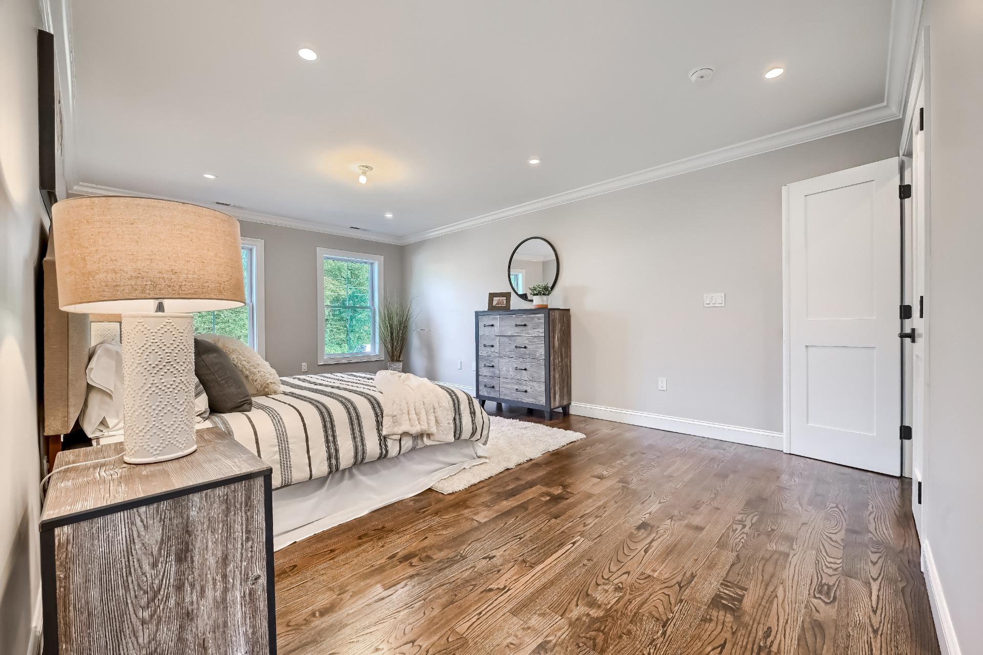 A bedroom with hardwood floors , a bed , dresser , lamp and mirror.
