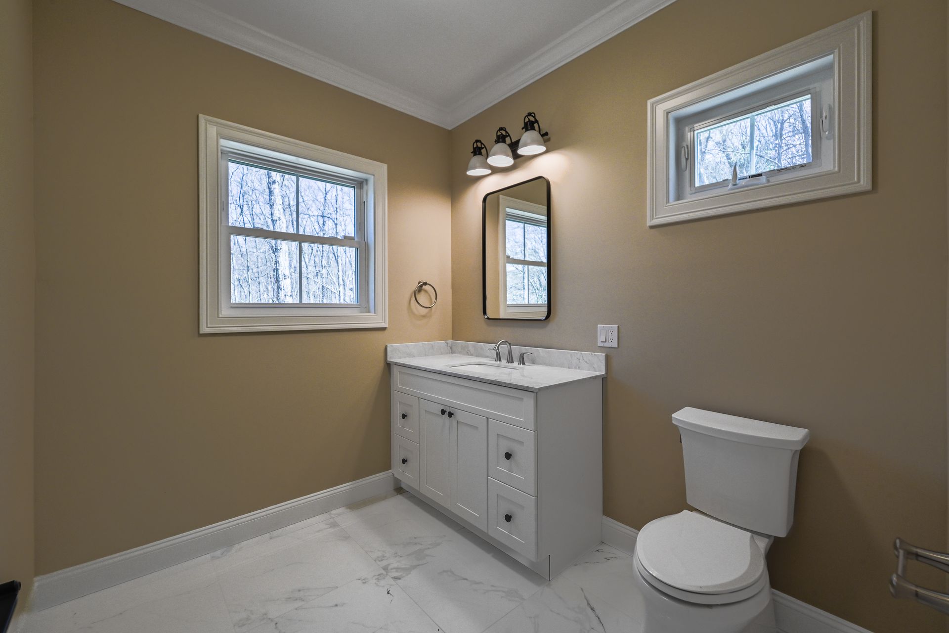 A bathroom with a sink , toilet , mirror and two windows.