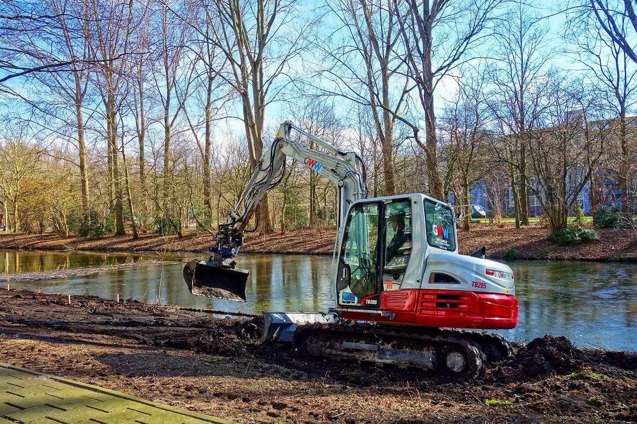 Excavator on muddy bank of a pond, scooping up soil. Trees line the water's edge on a sunny day.