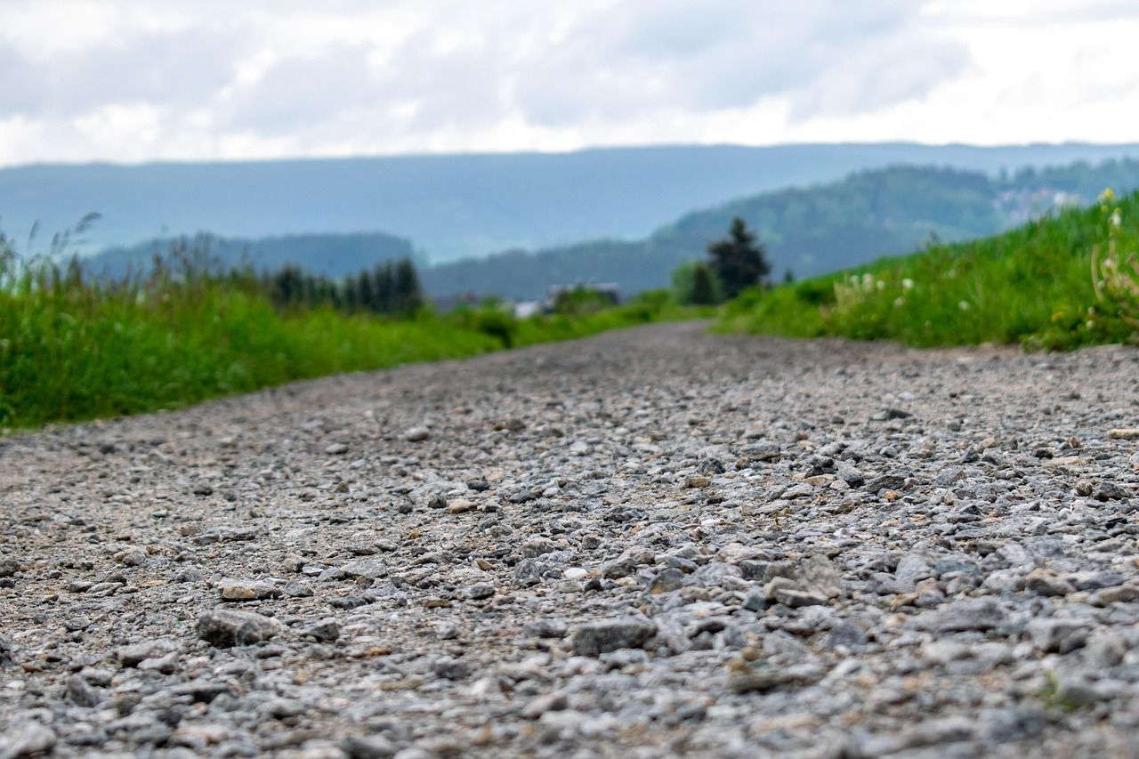 Gravel path through green field, leading toward distant hills under cloudy sky.