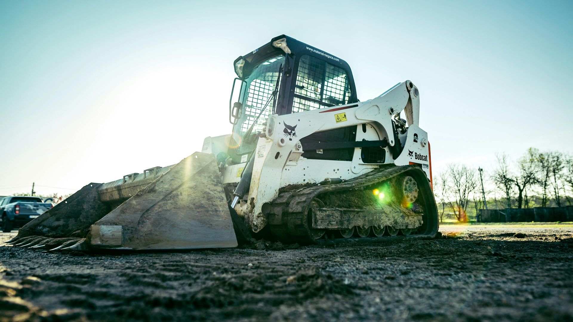 White Bobcat skid-steer loader on tracks, parked in a dirt lot with a pile of dirt in front. Sunlight shines.