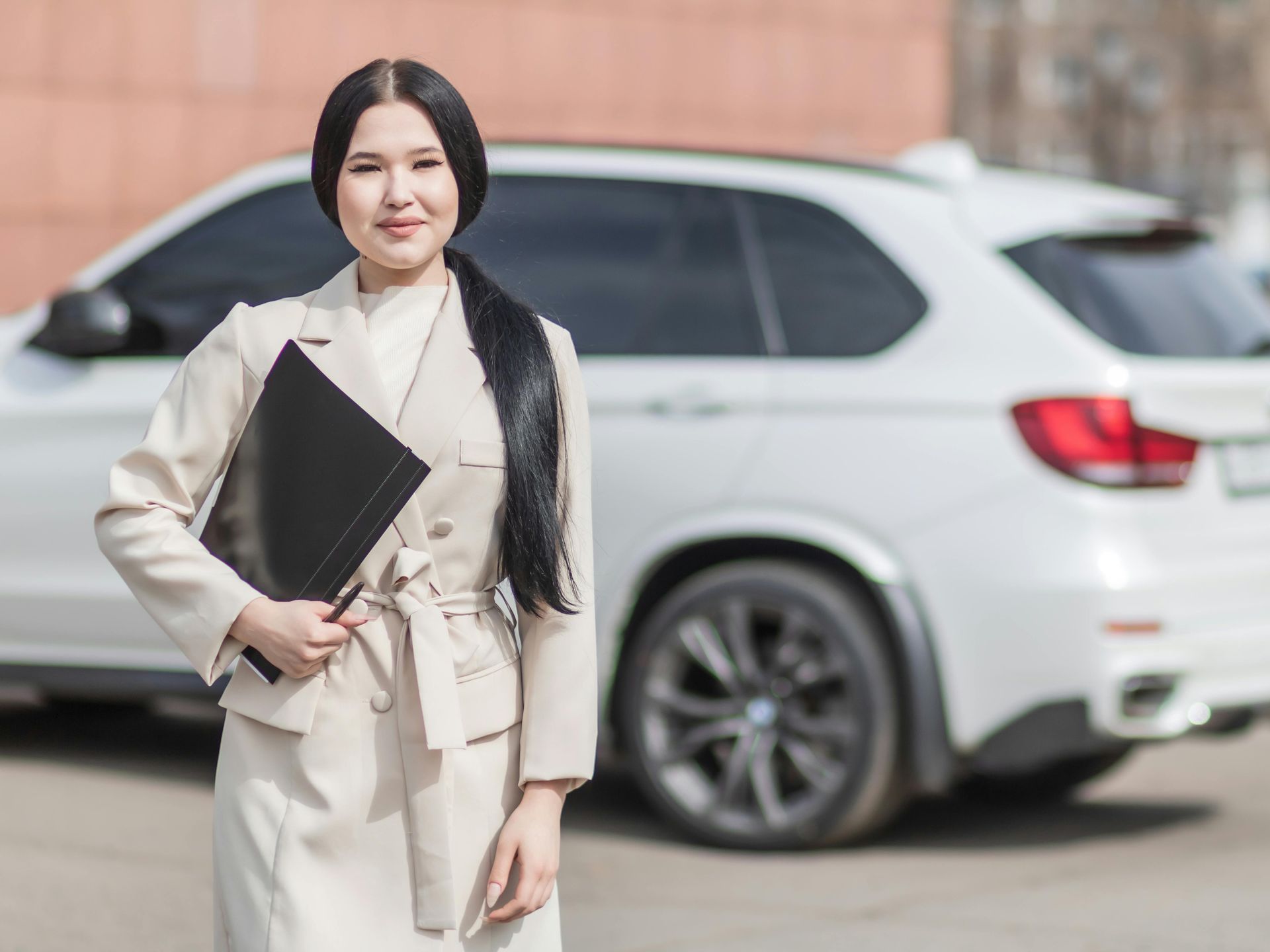 A woman is standing in front of a white car holding a folder.