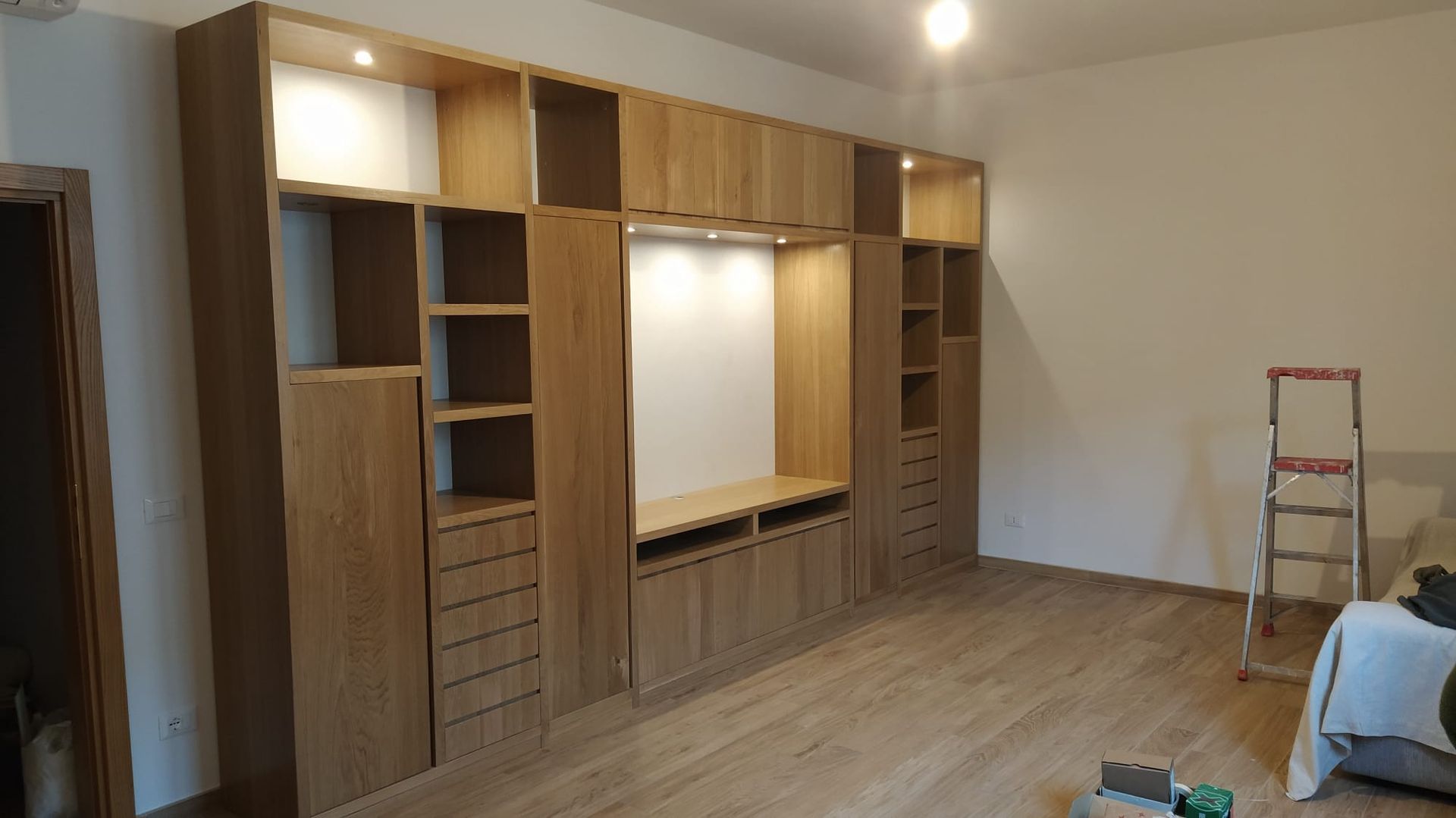 Mobile TV in legno con armadi, ripiani e cassetti. Lampadari installati. Pavimento e pareti in legno chiaro.