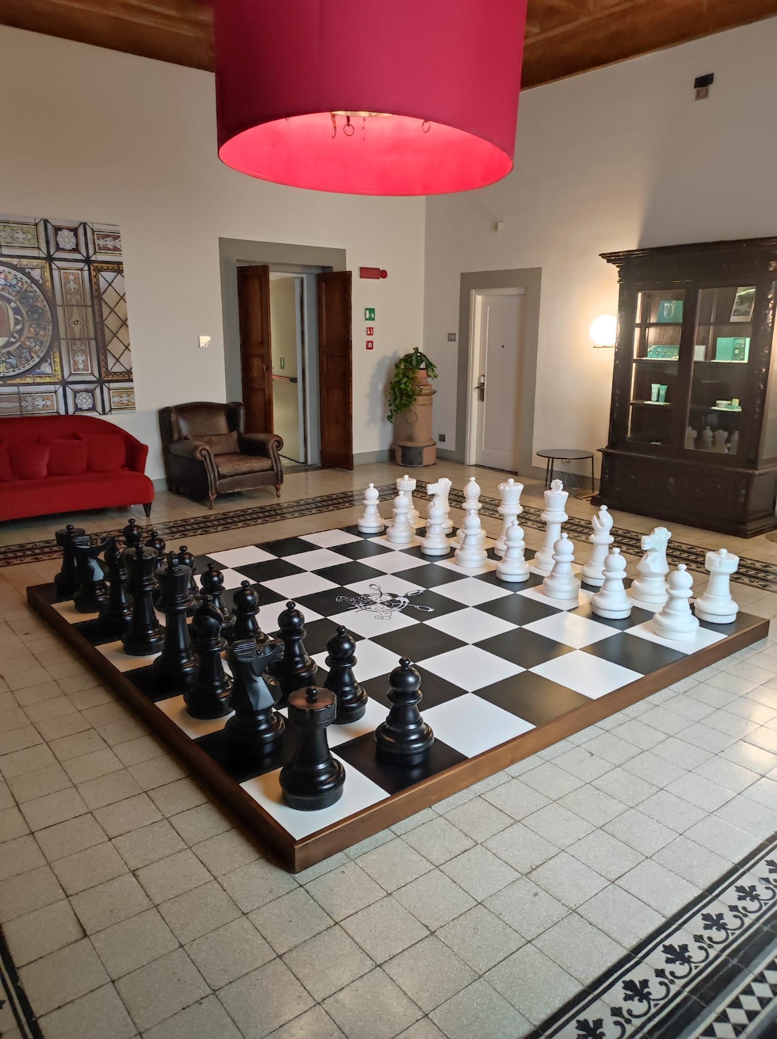 Scacchiera gigante all'interno con pezzi bianchi e neri. Lampada rossa sospesa in alto.