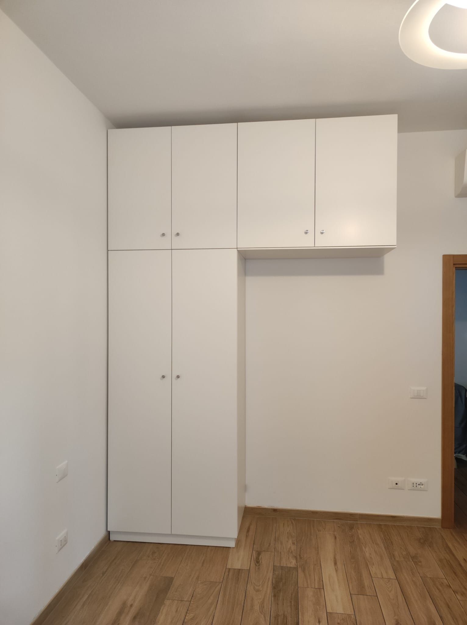 Mobili bianchi su una parete, sopra un pavimento in legno. I mobili includono unità alte e basse.