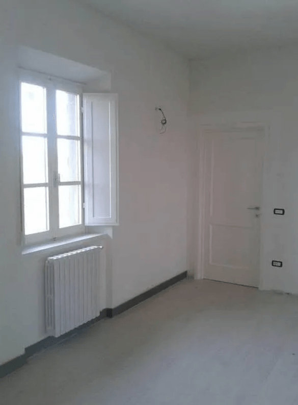 Stanza bianca e vuota con una finestra, un termosifone e una porta.