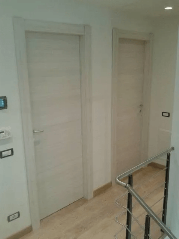 Due porte chiare in un corridoio bianco. Pavimento in legno, ringhiera in metallo e interruttori della luce a parete.