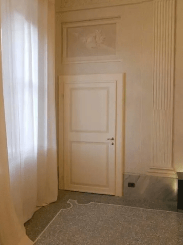 Stanza luminosa con porta bianca, tenda trasparente e modanature decorative. Pareti e moquette beige chiaro.