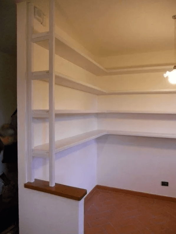 Mensole bianche a muro in un angolo, con ripiano in legno. Pavimento e pareti marrone chiaro.