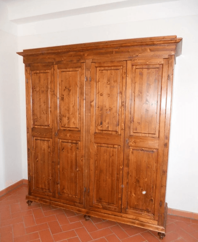 Armadio in legno a quattro ante, tinto marrone, appoggiato a una parete bianca su un pavimento in piastrelle di terracotta.