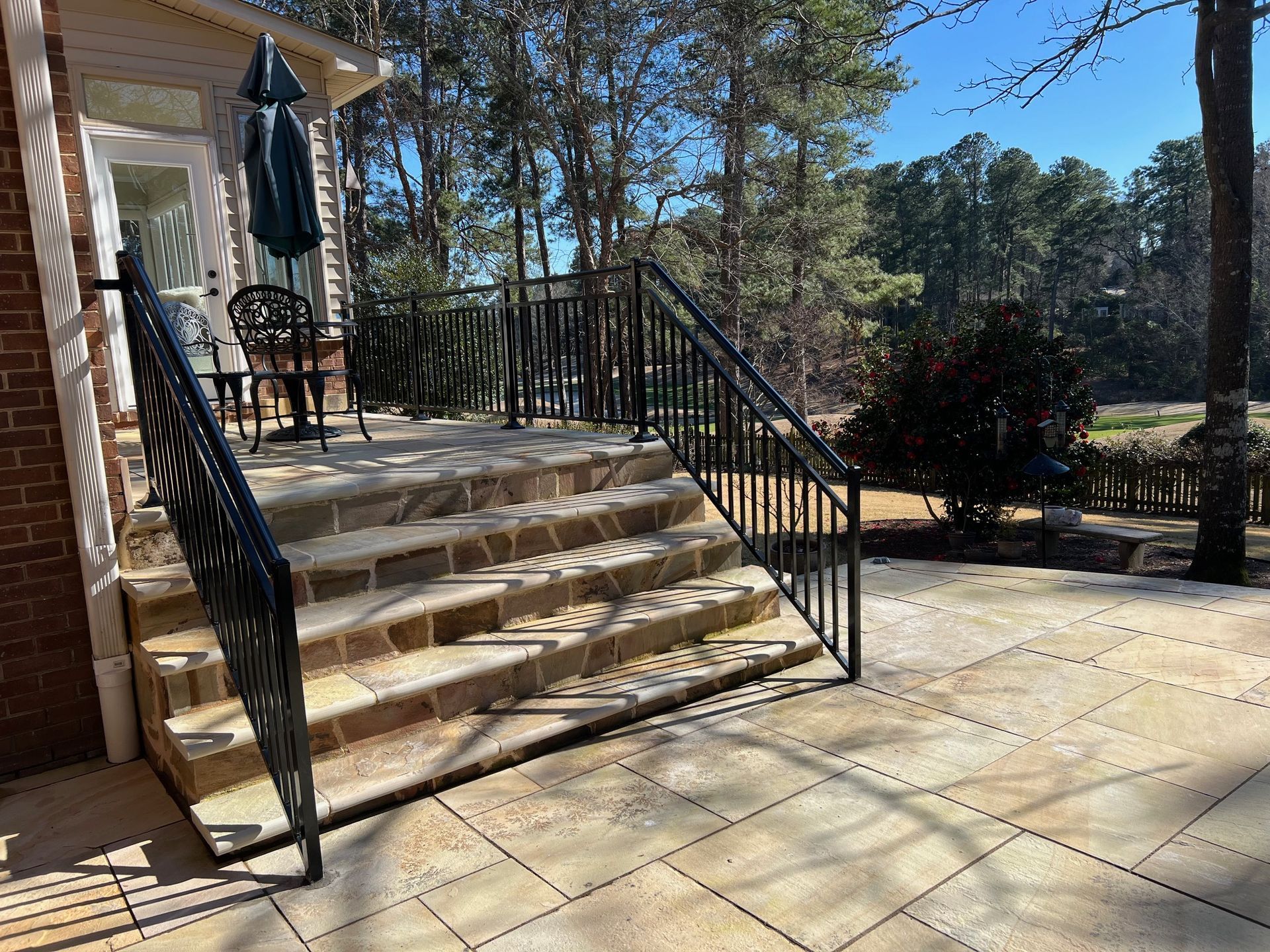 Stone steps lead to a patio with black railing and outdoor seating; trees in the background.