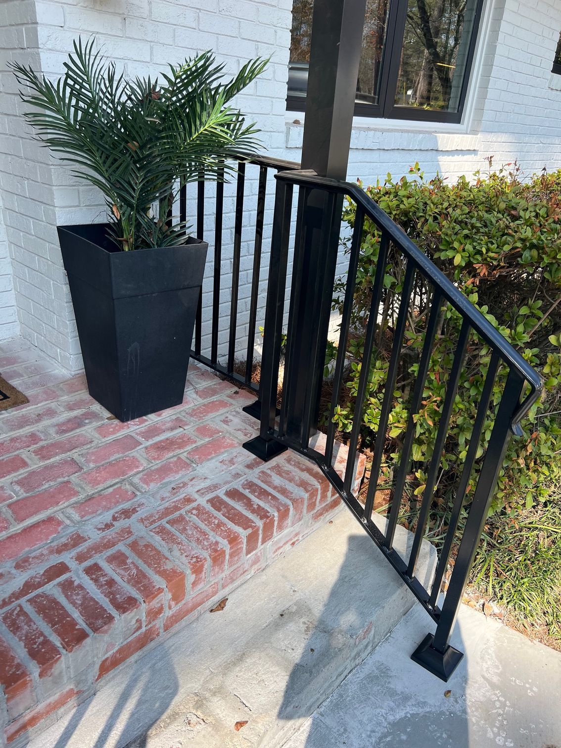 Black metal handrail on brick steps next to a potted plant and a bush.