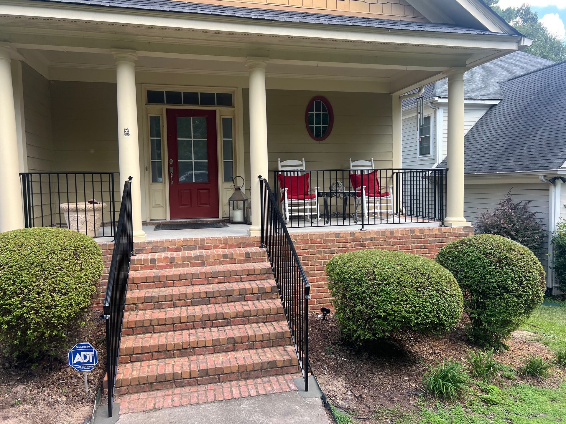 House with red door, porch, brick steps, black railing, and bushes.
