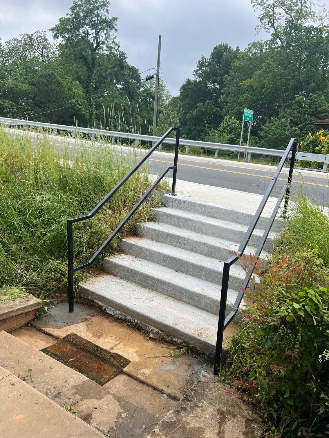 Concrete stairs with black handrails leading up to a road. Green foliage surrounds the stairs.