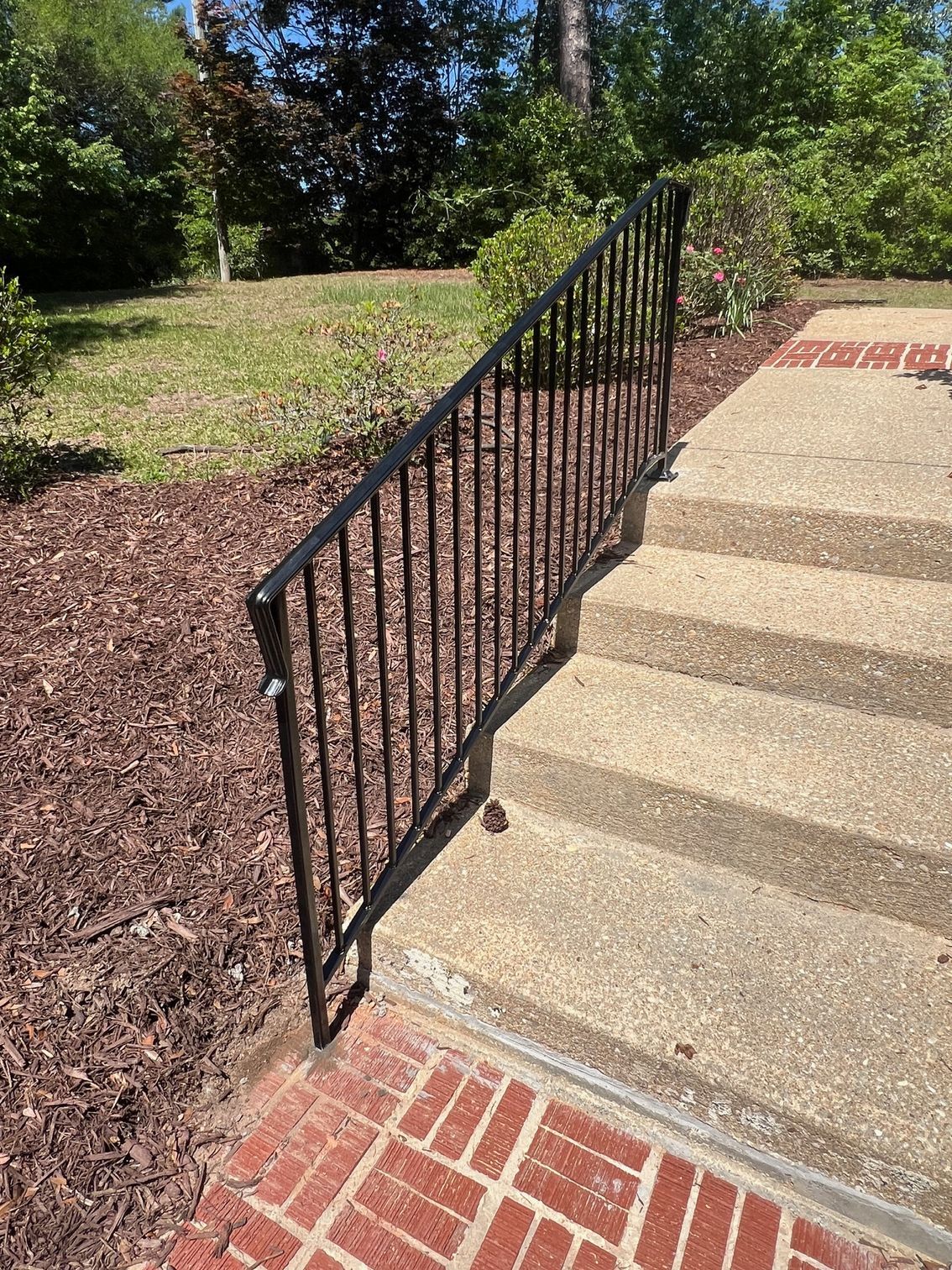 Black metal handrail on concrete steps, bordered by brick and mulch.