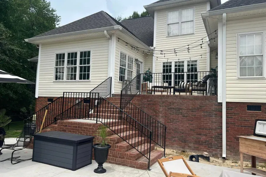 Back of a two-story beige house with brick foundation and a black railing staircase leading to a patio.