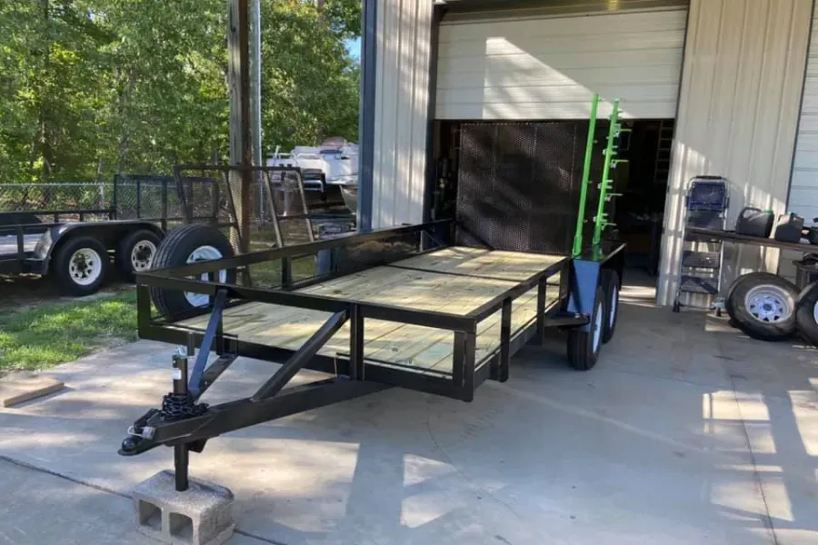 Black utility trailer parked in front of a garage, wooden deck, green shrub and spare tire, blue sky.