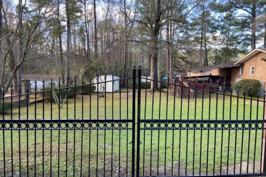 Black wrought iron gate overlooking a grassy yard with trees and a house in the background.