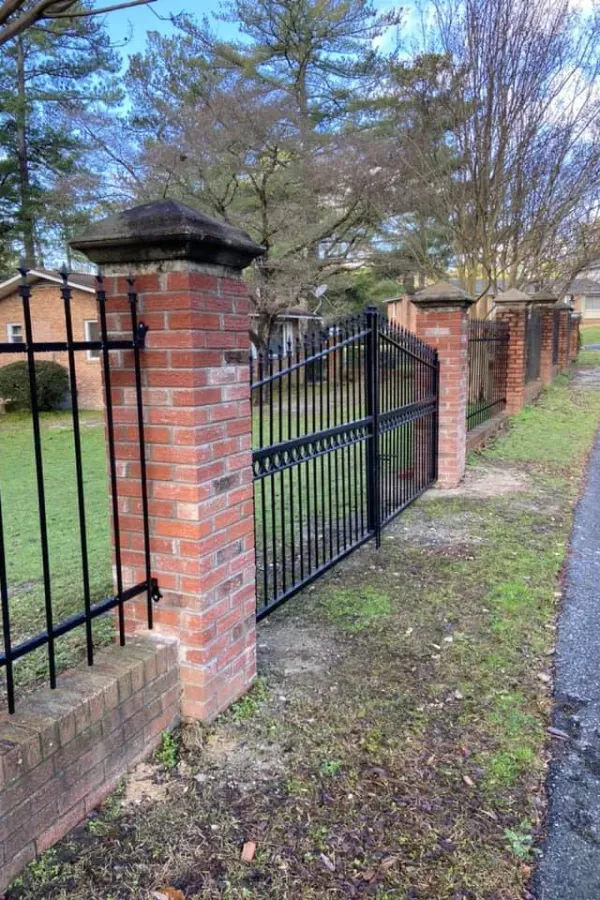 Black wrought iron fence with brick pillars and gate along a roadside.