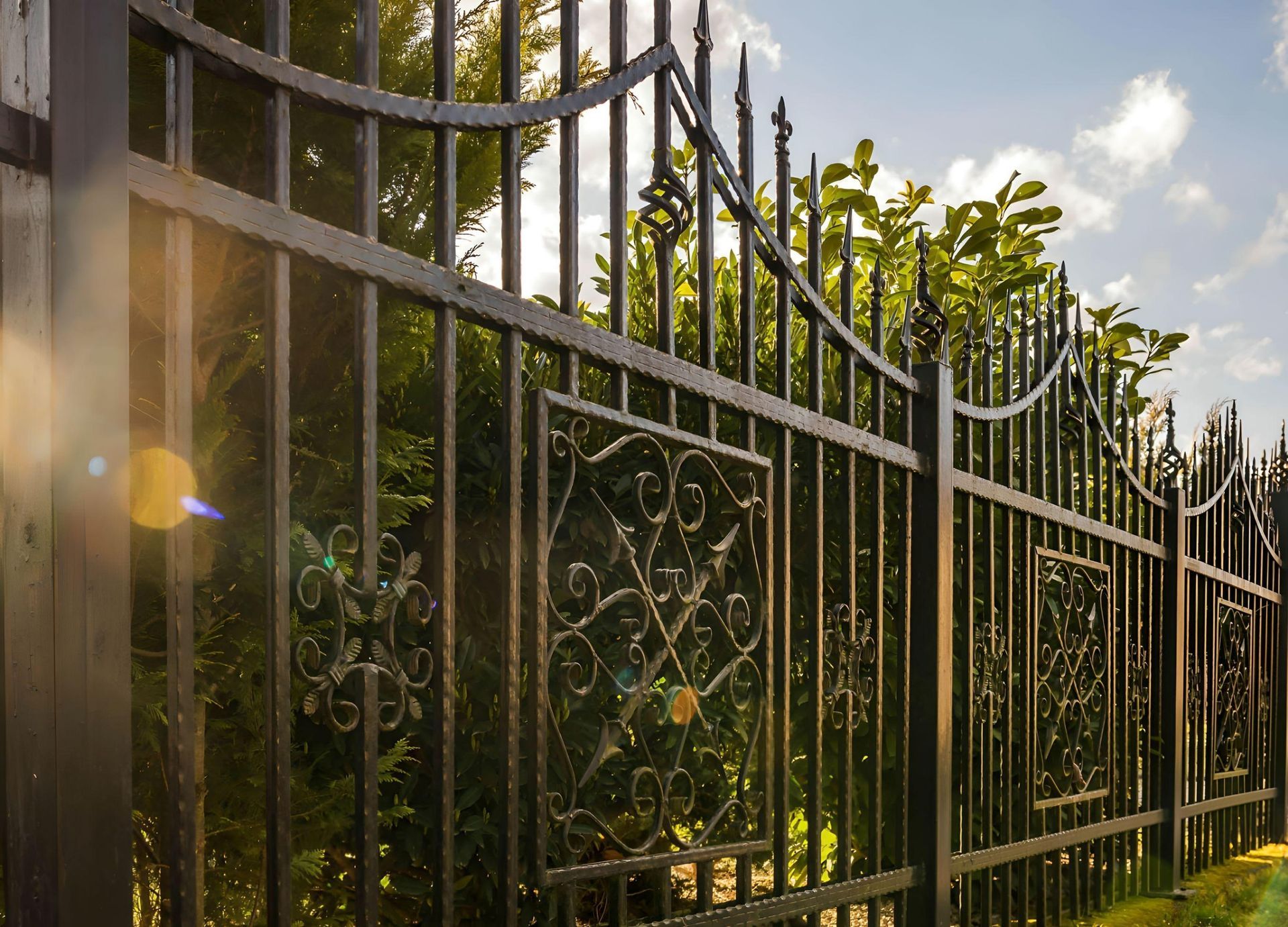 Black wrought-iron fence with decorative panels, set against a backdrop of green trees and a bright sky.