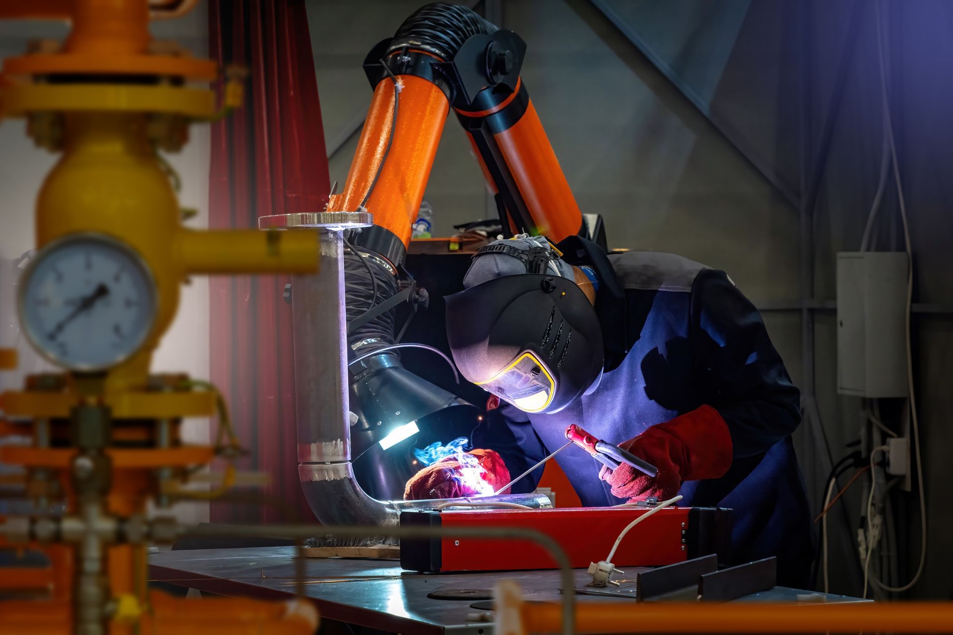 Welder in protective gear welding metal pipe in industrial setting, orange ventilation.